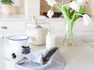 A calm and organised breakfast set up with a jar of oats, a ramekin of blueberries and milk in a jug. There is a linen napkin and a teaspoon. A vase of white tulips sets off the scene.