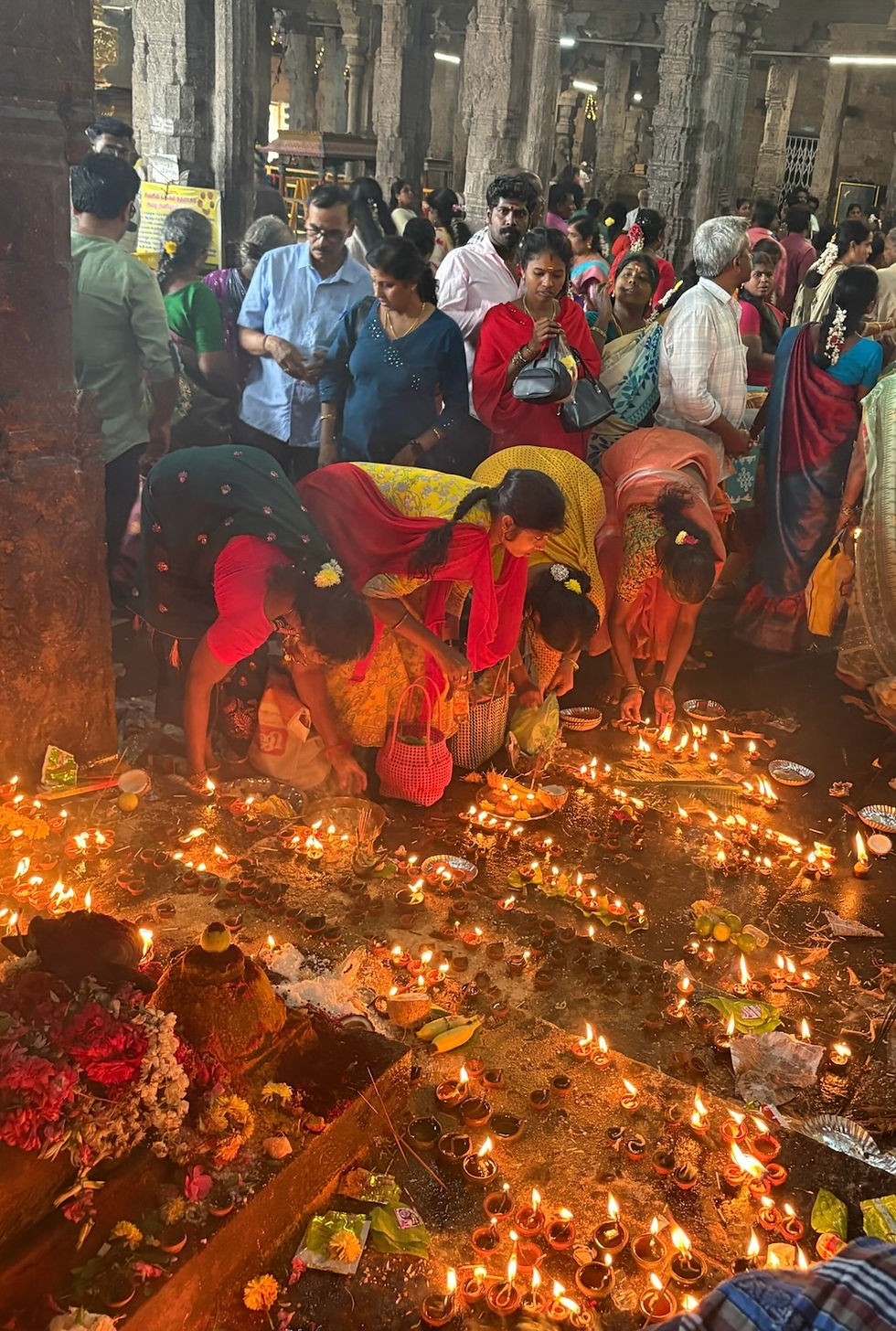 People gathered in a temple lighting clay lamps on the floor, creating a warm glow. Vibrant saris and flowers add color. Busy, devotional atmosphere.