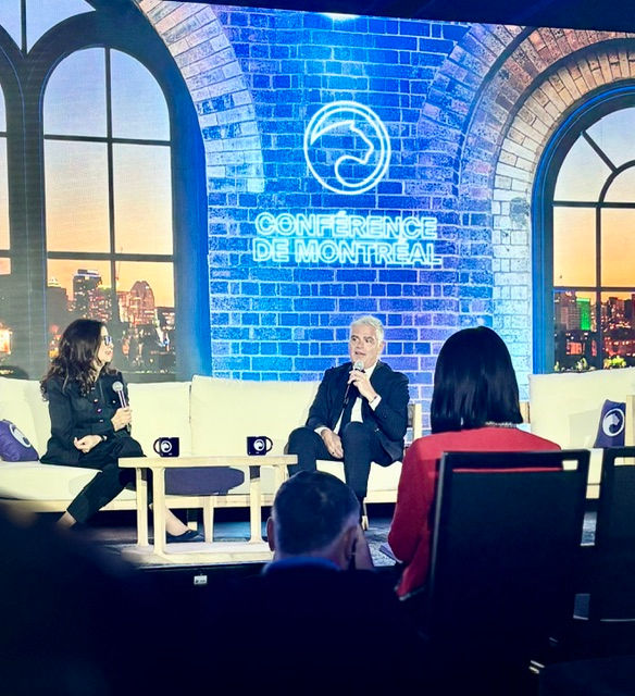 Panel discussion onstage at Conference de Montreal, two speakers on white sofas before a blue brick backdrop and city-window set.
