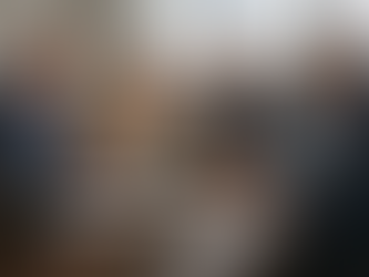 Four people in business attire having a discussion at a conference table with a laptop, papers, and a large window in the background.