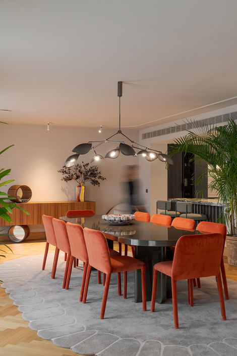 Contemporary dining room in a residential project in Portugal, with a polished oval table, warm-toned upholstered chairs, an organic-shaped rug and sculptural lighting, seamlessly connected to the living area with natural plants.