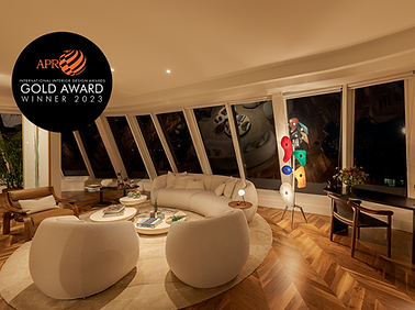 Sala de estar elegante e aconchegante na cobertura, com um sofá branco curvo, duas poltronas arredondadas e mesas de centro circulares dispostas sobre um tapete circular bege sobre piso de madeira em espinha de peixe. Uma luminária de chão escultural multicolorida fica ao lado de uma escrivaninha com um arranjo floral e duas cadeiras de madeira. Janelas angulares do chão ao teto emolduram uma vista noturna da cidade. No canto superior esquerdo, um emblema circular preto exibe o logotipo do APR International Interior Design Awards com o texto: "GOLD AWARD WINNER 2023".