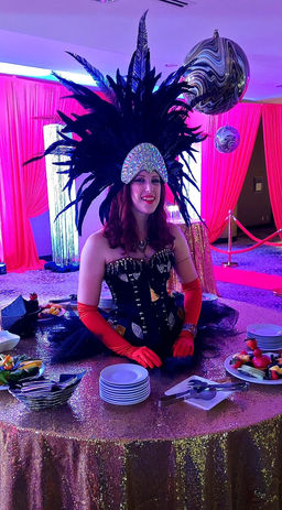A woman wearing a black corset, red gloves, and a black feather headdress, is inside of a strolling table. The table is decorated in gold and black.