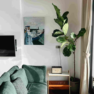 Interior de una sala de estar con un sofá verde, flores en la habitación, televisión, altavoz y cortinas, acompañado de un cuadro de un marinero con un bote en la pared.
