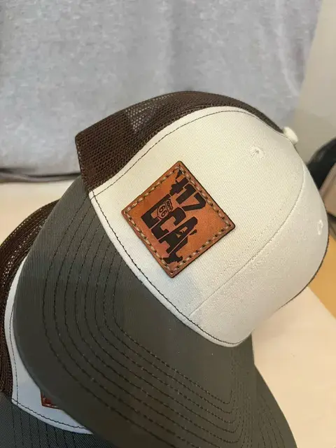 Thumbnail: Close-up of a white and gray trucker hat with brown mesh backing and a bold leather "417 Local" Route 66 patch stitched on th