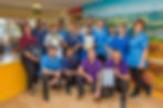 A group of care home staff in blue and purple uniforms holding certificates, smiling. Background shows a scenic landscape mural.