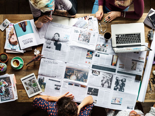 Colleagues working around a desk covered in newspaper spread and laptops and tablet computers