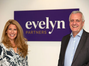 Two staff members in front of a purple "Evelyn Partners" sign in it's new branding style. The woman wears a floral top, and the man wears a striped shirt and blazer.