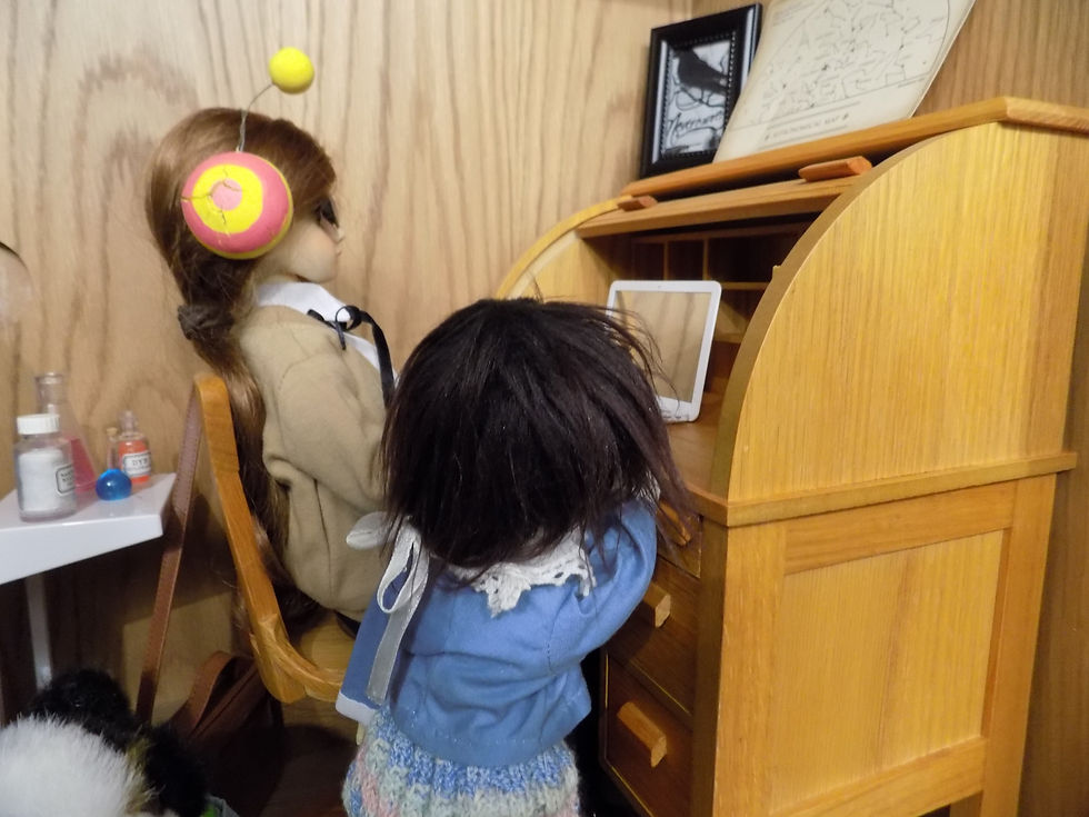 Aster, an adolescent doll wearing an "alien" style headband sits at a wooden desk, watching a laptop. A child doll with her back to the viewer, Nurit, stands next to the adolescent and watches the laptop as well. At the bottom left of the photo is a dog doll.