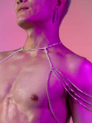 Topless man wearing a shoulder body jewelry. The piece is made of three flowing stainless steel and crystal chains, covering his shoulders and wrapping around his neck. 