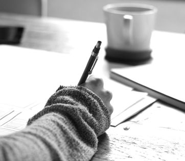 A black and white shot of a desk covered in papers, a mug in the background, a person's hand writing handwritten notes