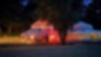Illuminated yurts with strings of lights in a dark wooded area. A person in the distance by a path lit with lanterns, creating a warm glow.