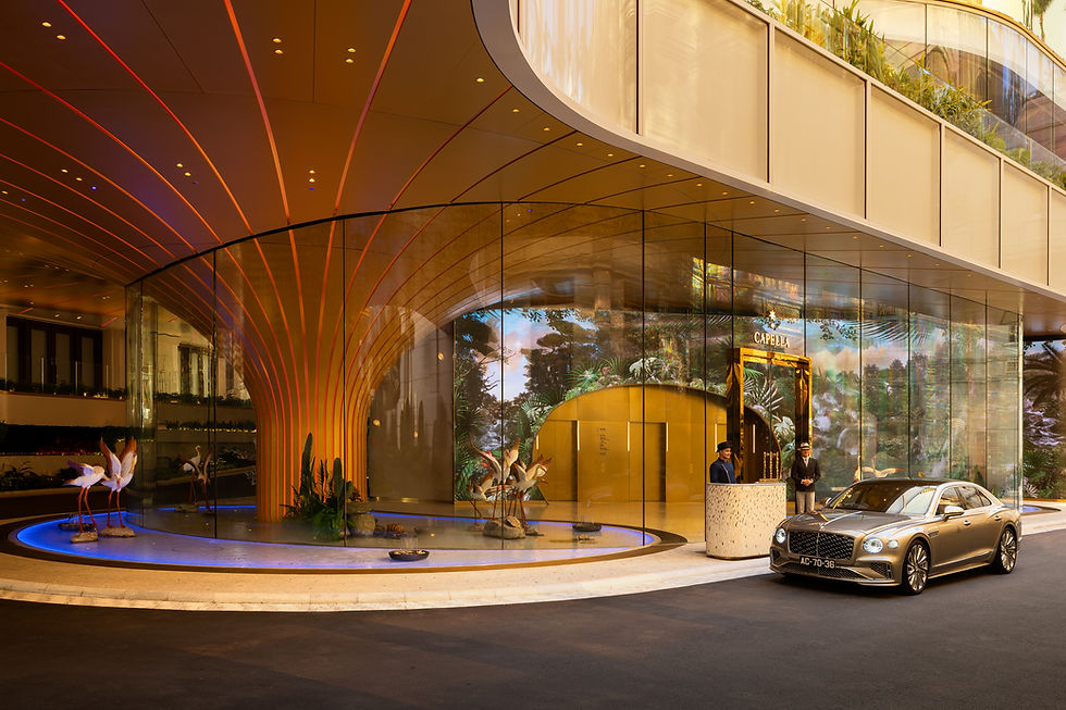 A luxury car is parked outside a modern, glass-fronted building with curved architecture and a covered entrance, where two staff members stand by the door.