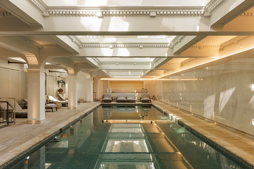Indoor pool with a serene ambiance, surrounded by lounge chairs and neutral tones. Sunlight filters through a glass ceiling, creating reflections.