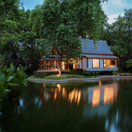 Serene evening scene of a traditional house by a pond, illuminated warmly. Surrounded by lush trees and a peaceful waterfall, reflecting on water.