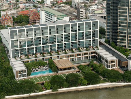 Aerial view of a modern hotel by a river, featuring a blue pool and green rooftop gardens. City buildings and a red-roofed temple nearby.