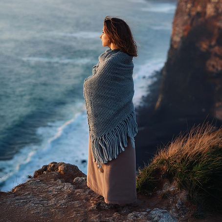 Woman in a gray shawl stands on a cliff edge, overlooking the ocean. Rugged coastline and waves create a contemplative mood.