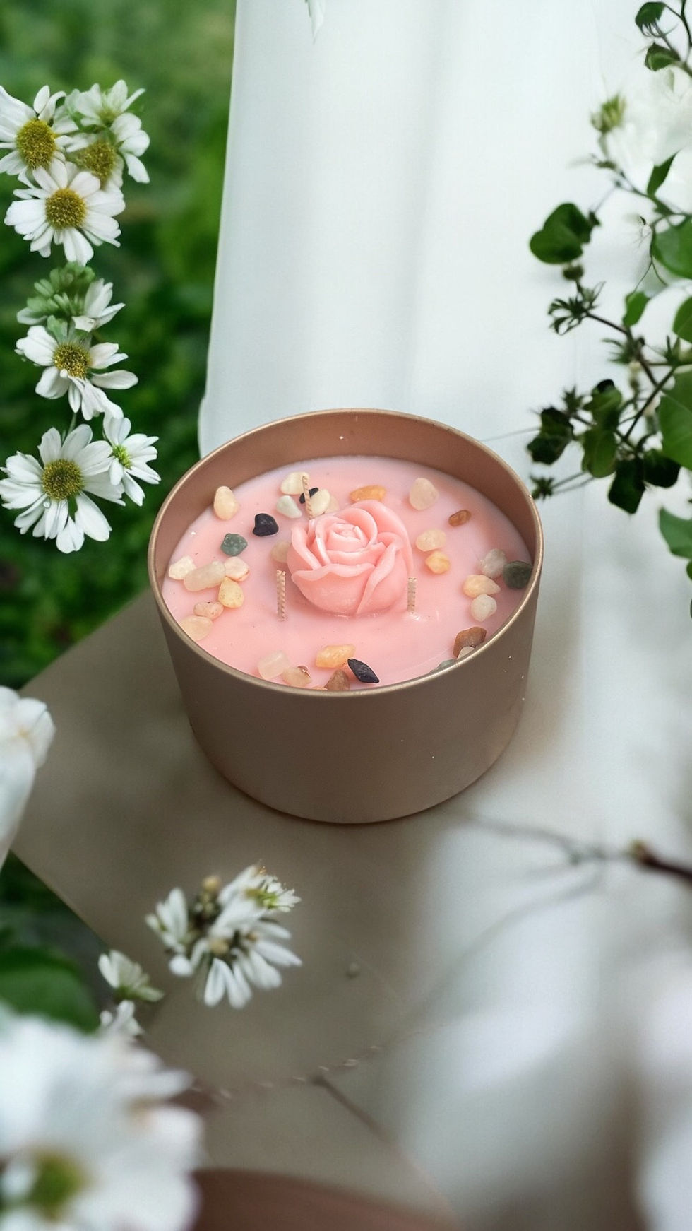 Pink Rose candle with beach stones in a champagne tin container sitting on a white background surrounded by white flowers.