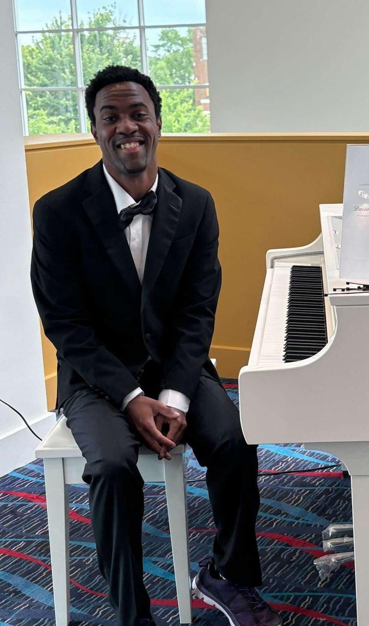 Darnell sitting on a piano bench in a tuxedo next to a white piano, smiling for the picture
