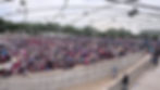 Large crowd seated in an outdoor amphitheater under a metal canopy, many holding umbrellas on an overcast day.