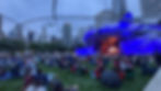 Crowd watches outdoor concert at dusk in a city park, blue-lit stage under curving canopy with skyline and Prudential tower.