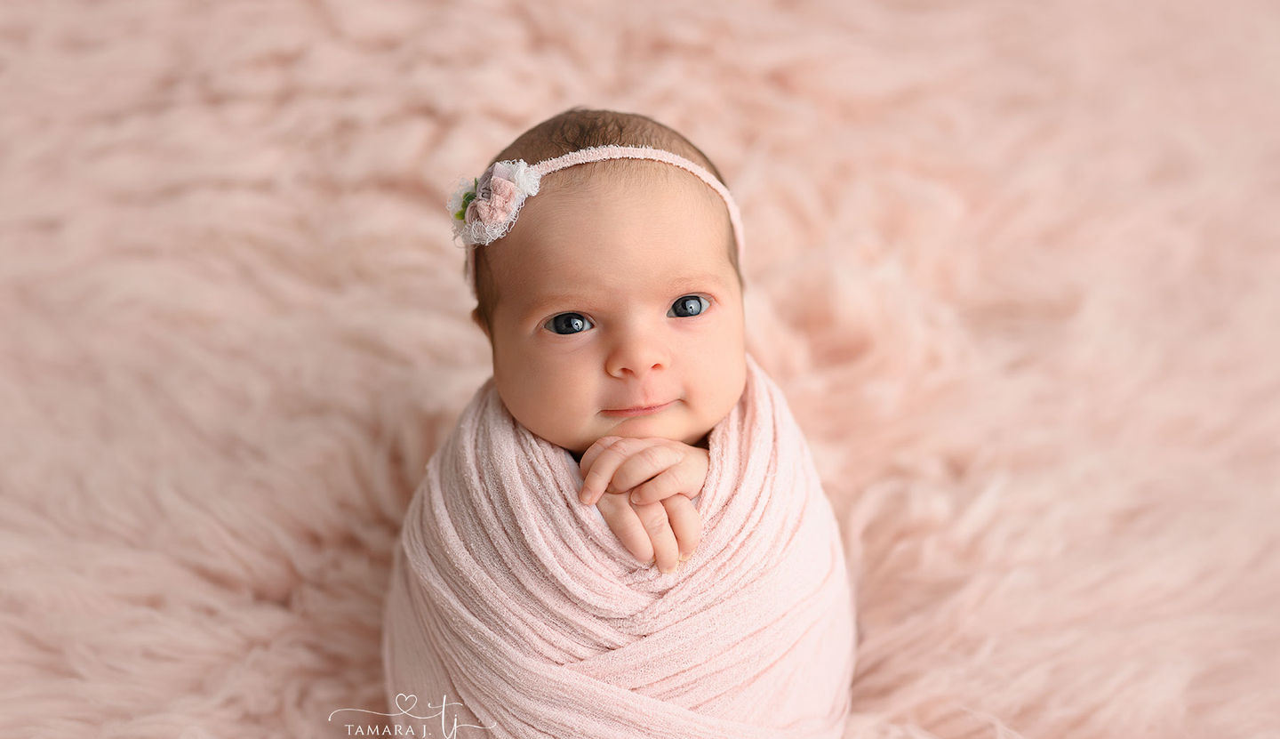 Newborn baby girl in pink wrap with floral headband, posed on soft pink fur by Calgary newborn photographer Tamara J. Photography