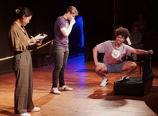 three actors on stage standing around a suitcase