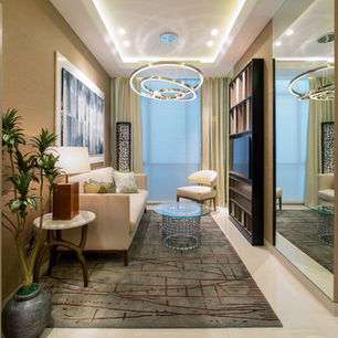 Area lounge yang hangat, nyaman, dan elegan, menampilkan sofa berwarna netral, karpet bermotif, pencahayaan geometris modern, serta jendela besar dengan tirai tipis (sheer curtains)