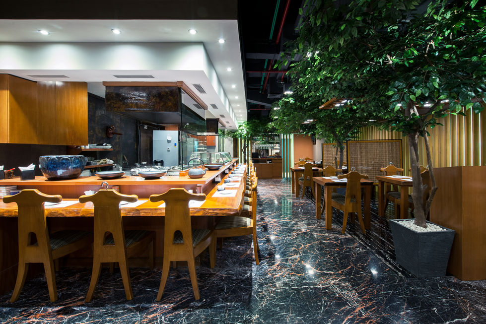 Japanese restaurant interior showing a long wooden sushi counter with chairs, polished marble floors, and an open kitchen area.