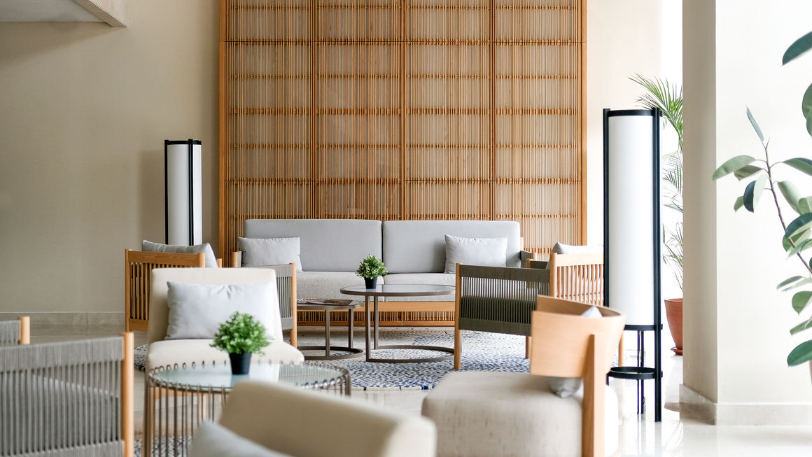 Hotel lobby seating area featuring a grey sofa, wooden armchairs, tall cylinder lamps, and a wooden slat backdrop.