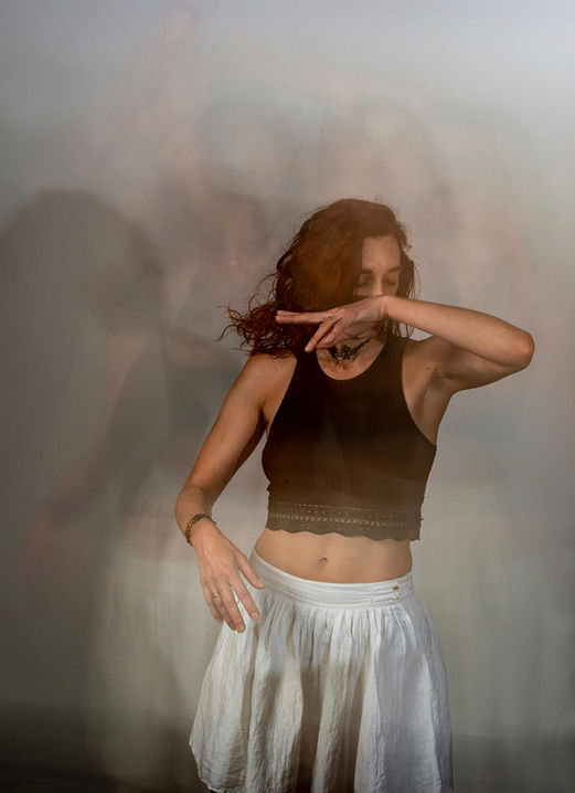 Portrait en mouvement d'une personne aux cheveux roux dansant ou effectuant une performance artistique. Elle porte un haut sombre et une jupe claire, et lève un bras pour cacher partiellement son visage. L'arrière-plan est flou avec des traces de mouvement, créant un effet onirique ou d'écho visuel qui suggère la fluidité et la vitesse du mouvemen