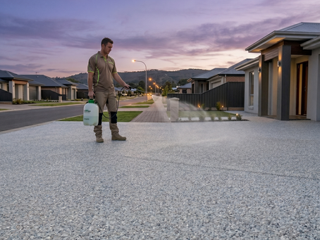 A professional tradesman in workwear standing on a wide residential driveway, using a manual pump pack pressure sprayer to evenly apply a clear sealer across a clean, light-grey exposed aggregate concrete surface. The entire driveway maintains its beautiful, true-to-life natural stone appearance without any artificial dark or brown discoloration, set against a modern Australian suburban house backdrop during sunset.