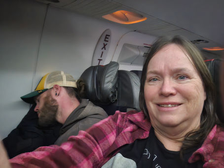 A smiling passenger takes a selfie on an airplane while the person beside her sleeps against the window near the exit row.