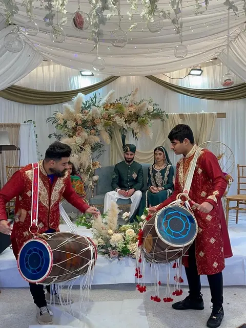 Traditional Pakistani wedding ceremony featuring a bride and groom in elegant attire, with a stunning floral backdrop of pampas grass and roses, accompanied by dhol drummers in red and gold outfits