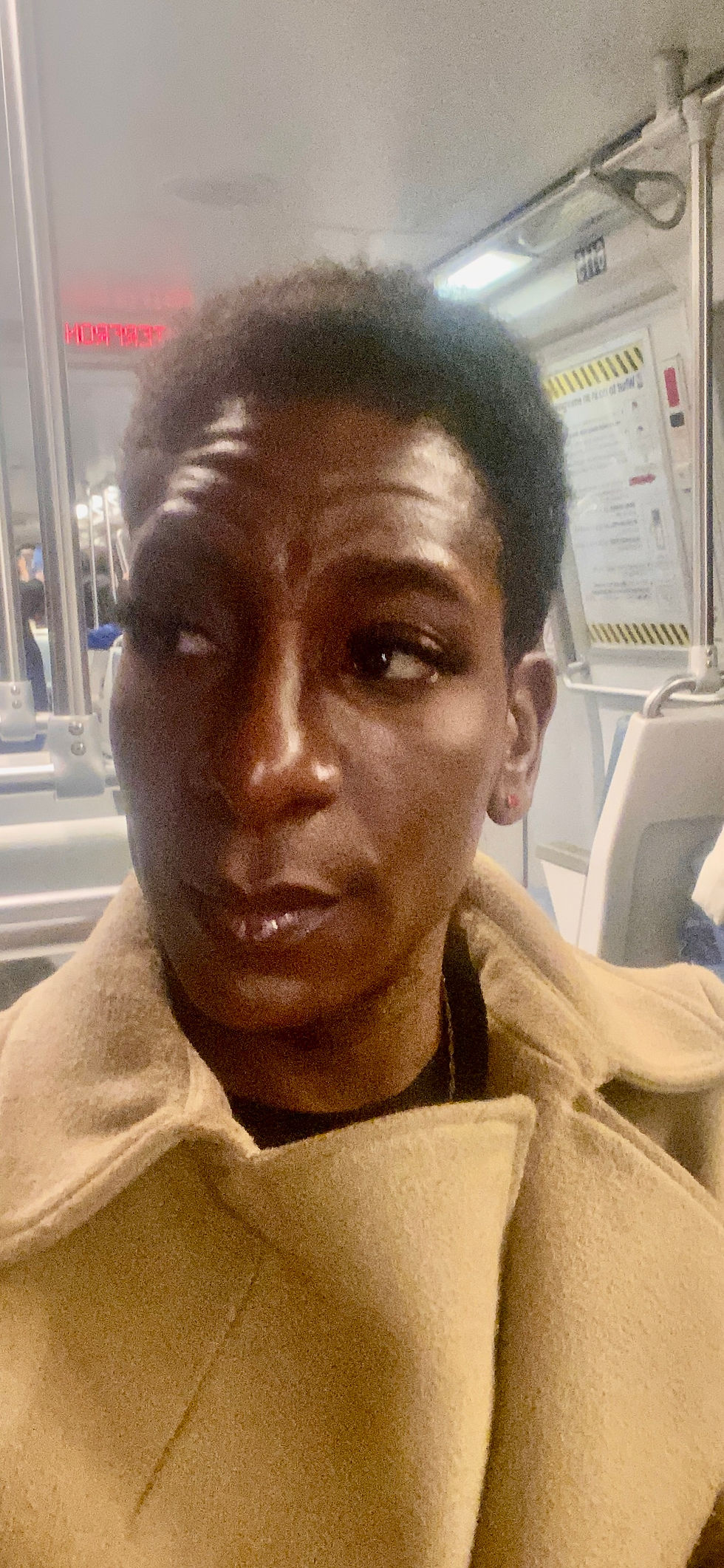 Yannick is seated on the train. She is wearing a tan coat. She has a short hair cut and is looking towards her left