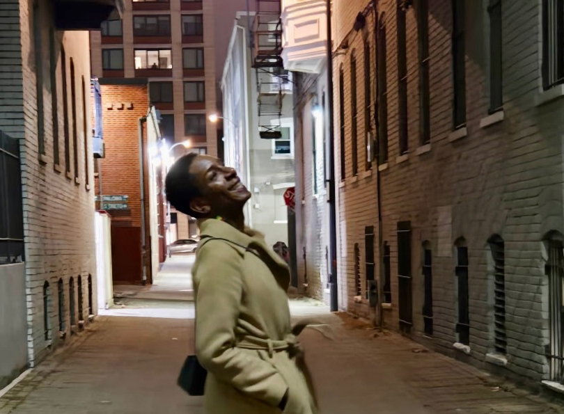 Yannick looking to the right in a lighted alley way. She is wearing a tan coat with her hands in the pocket. She has a small pocket book on her left side. Yannick is wearing Black boots.