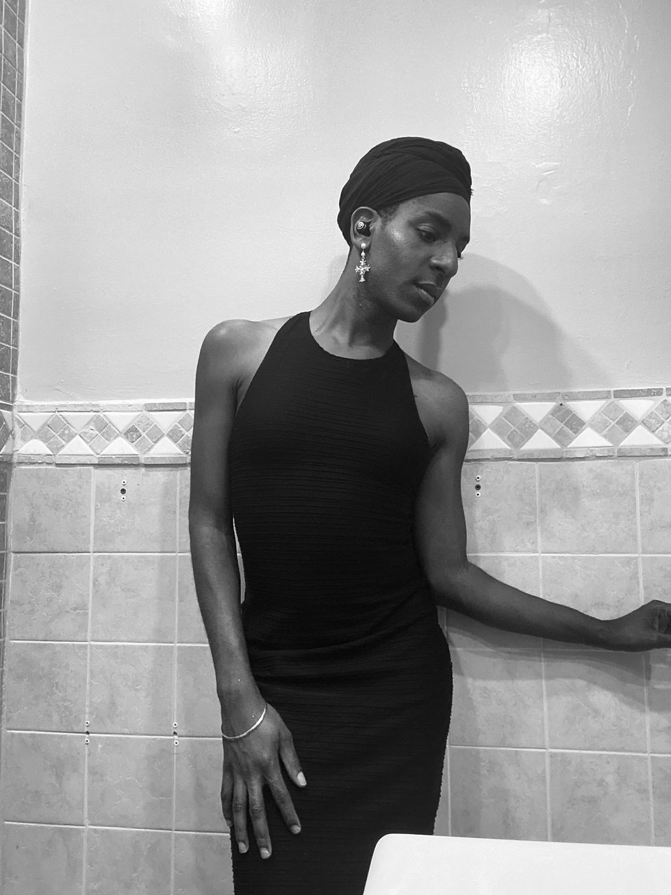 Yannick is pictured in a monochrome filter. She is wearing a Black head wrap with cross earrings. Yannick is wearing a long Black Fitted dress that
sleeveless. She has on a silver bracelet on her right hand.