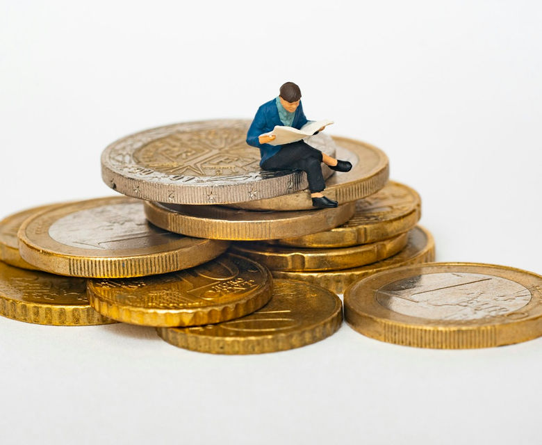Miniature person sitting on stack of coins reading newspaper