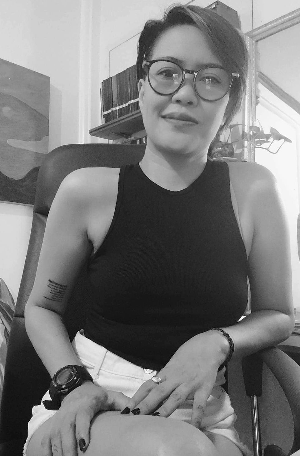 Smiling person with short hair and glasses sits on a chair. Wearing a black top and white shorts. Tattoo visible. Monochrome setting.
