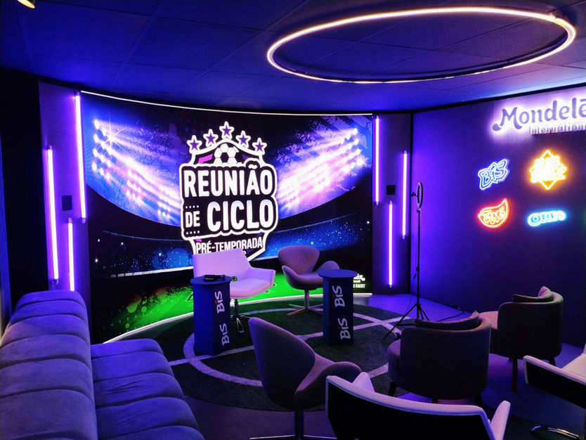 Ambiente intimista todo roxo, realizado para a mondeles. Ao fundo um painel escrito "Reunião de Cliclo" com uma luminária circular no teto e vários estofados pelo ambiente.