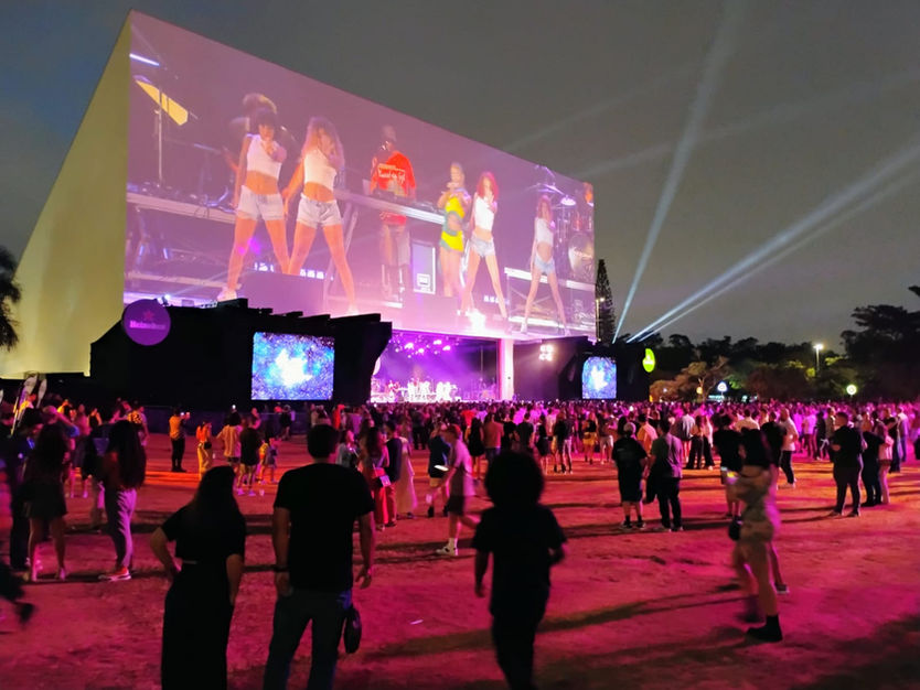 Espaço aberto com uma projeção gigante em destaque, um palco logo abaixo da projeção e um público massivo ocupando o espaço
