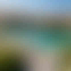 Aerial view of a lakeside village with people strolling. Clear turquoise water, rustic buildings, and mountains under a bright blue sky.