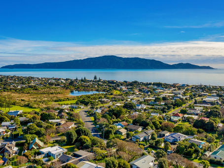 Aerial view of a seaside town with green rooftops and lush vegetation. Calm blue sea and distant mountains of Kapiti Island under a clear blue sky.