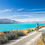 Thumbnail: Cyclists ride along a gravel path beside a turquoise lake under a clear blue sky.