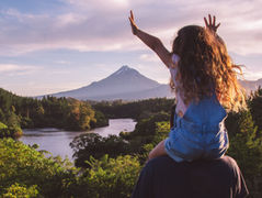 A child in blue overalls sits on a shoulder, arms raised joyfully. They overlook a lush forest and a snow-capped Mount Taranaki under a pink sky.