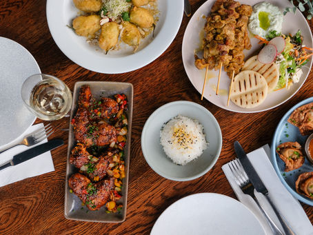 Various dishes on a wooden table, including fried appetizers, skewers, white rice, and a glass of wine, creating a warm dining atmosphere.