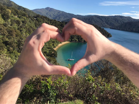 Hands form a heart shape framing a scenic view of a sailboat on a blue-green bay in the Marlborough Sounds, with lush green hills under a clear blue sky.
