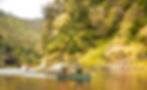 Two people paddle a green canoe on the Whanganui River, embarking on a classic New Zealand adventure amidst lush green hills.
