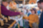 A woman in a yellow sweater smiles as she exchanges money with a vendor at an outdoor market stall in Radiant Nelson.