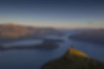 Thumbnail: A person in a red jacket stands on a grassy peak, overlooking the serene Lake Wanaka with several small islands.